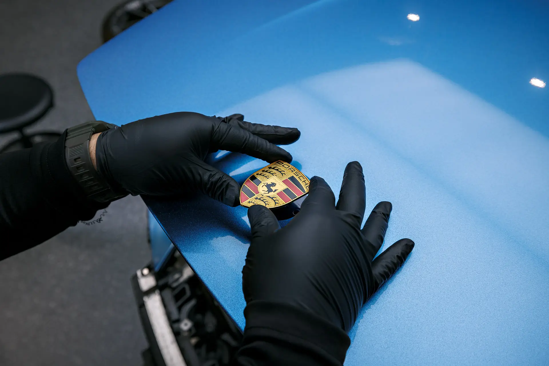 Gloved hands holding a Porsche crest over a blue work surface