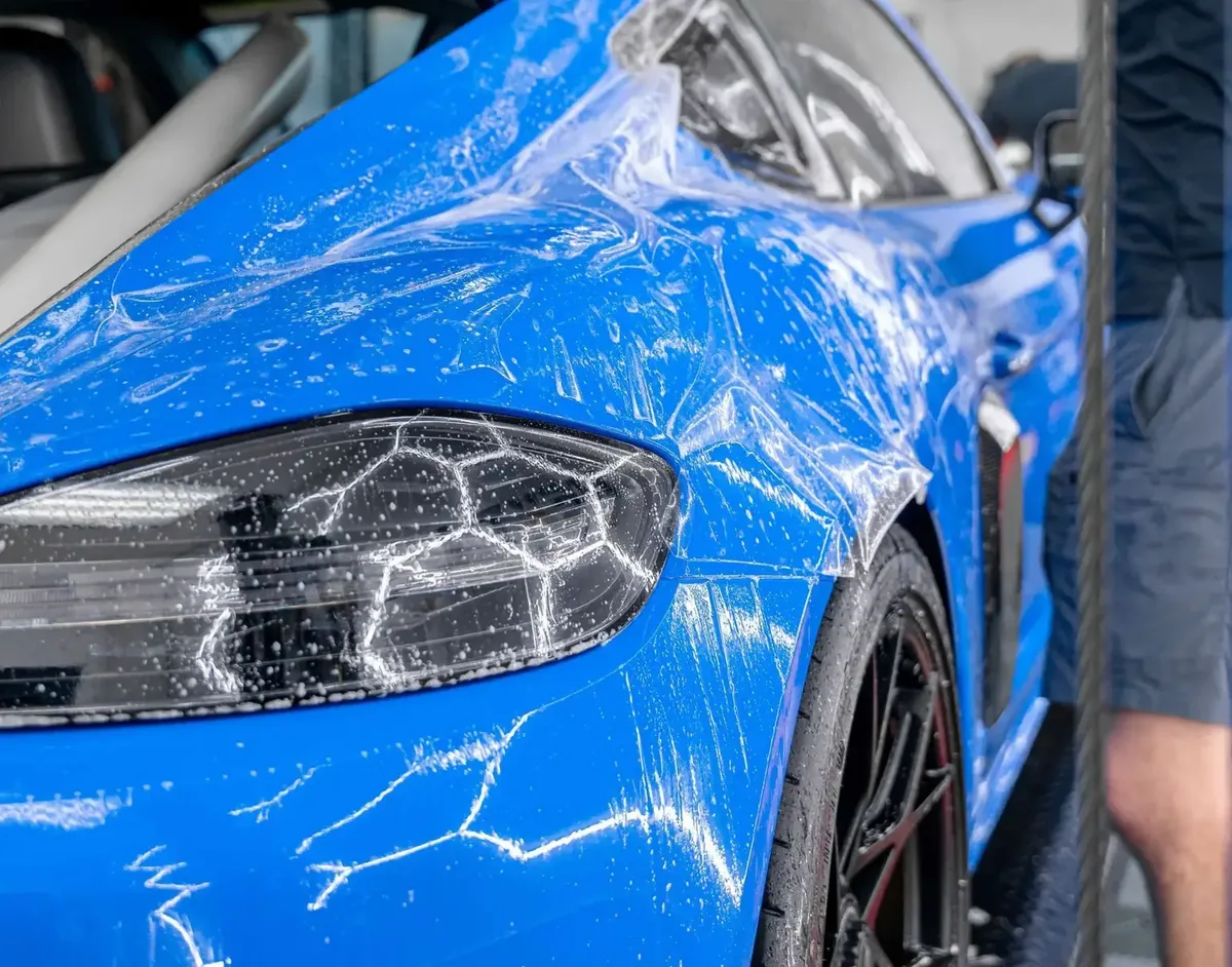 Front corner of a blue sports car with wet paint protection film laid over the headlight and front wing, slip solution streaked across the surface