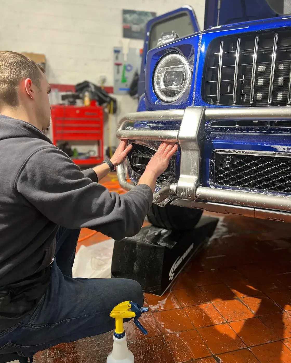 Technician crouched at the front of a blue Mercedes G-Class, fitting paint protection film around the bumper and brush guard, with a red tool chest behind him in the shop