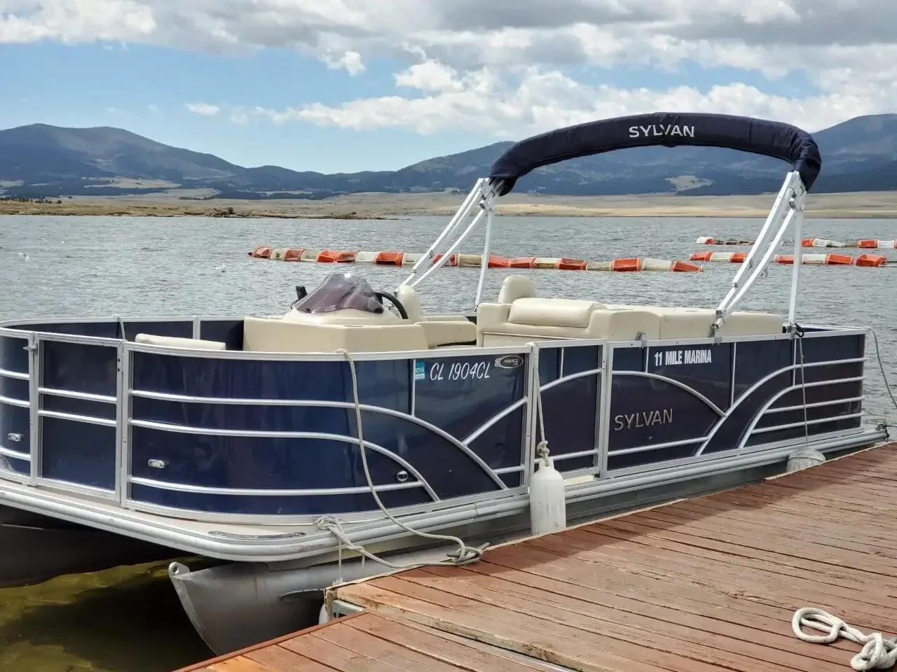Navy Sylvan pontoon boat tied to a wooden dock on a lake with mountains behind