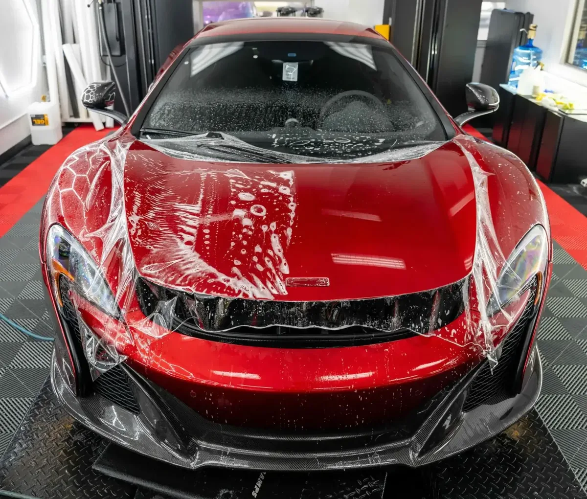 Red McLaren photographed head-on in the studio bay with wet paint protection film laid across the full nose, hood and front wings, slip solution pooled over the surface