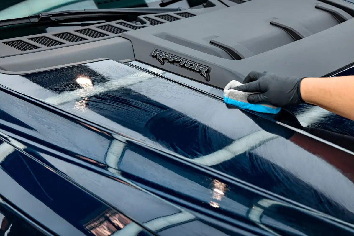 Gloved hand working a product across the hood of a dark blue Ford truck with a foam applicator block