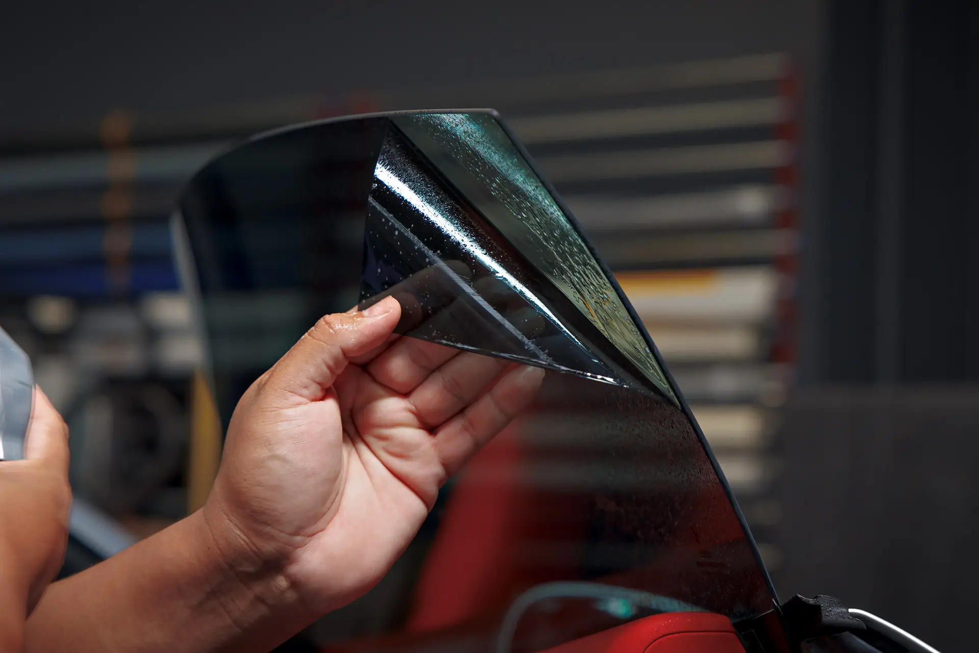 Hand holding a cut sheet of window tint film up to the light in front of racks of film rolls at ST6 Detailers