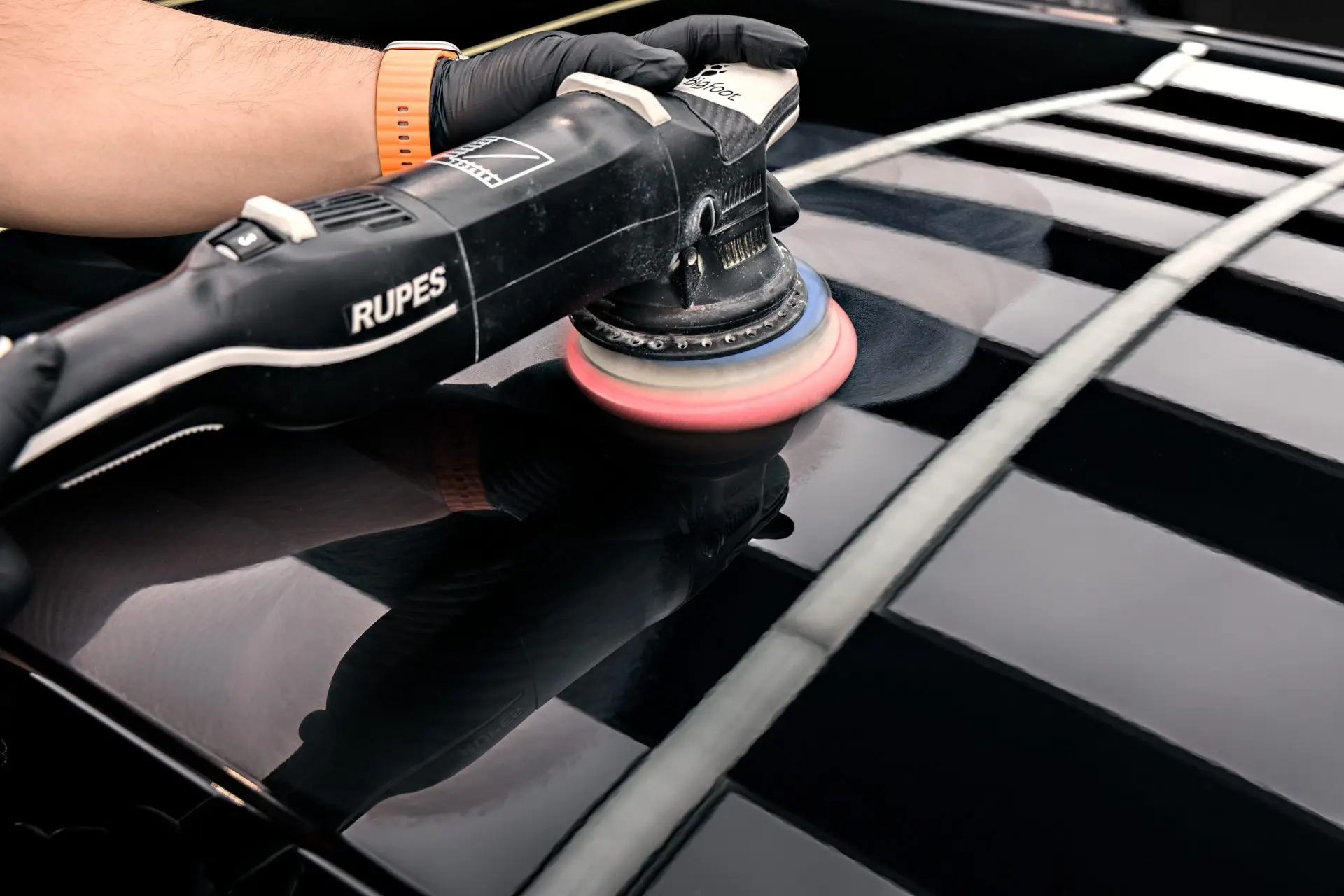 Close-up of a RUPES polisher with an orange pad working a black panel under strip lights