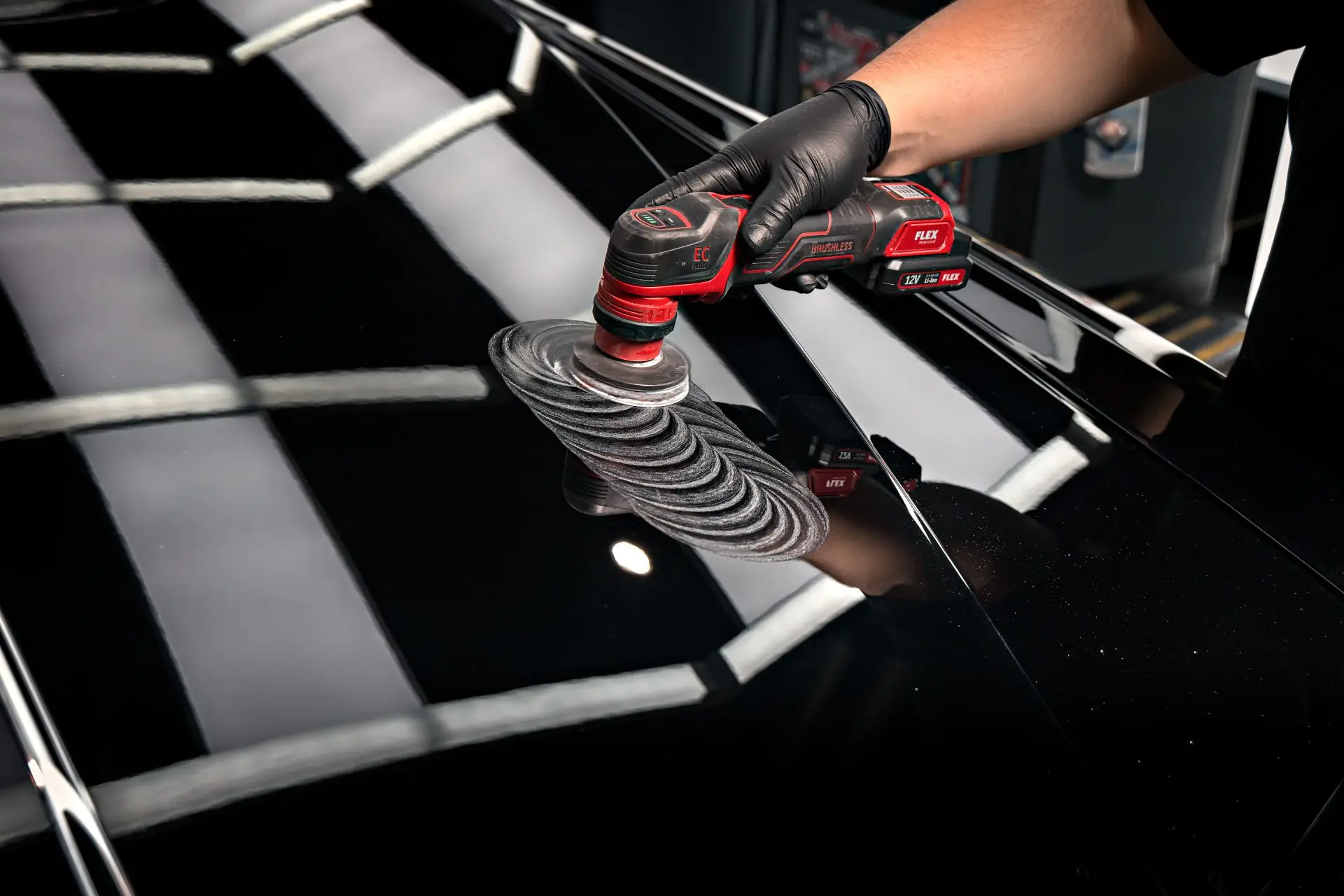 Gloved hand running a compact polisher across black Rolls-Royce Phantom paint under shop lights