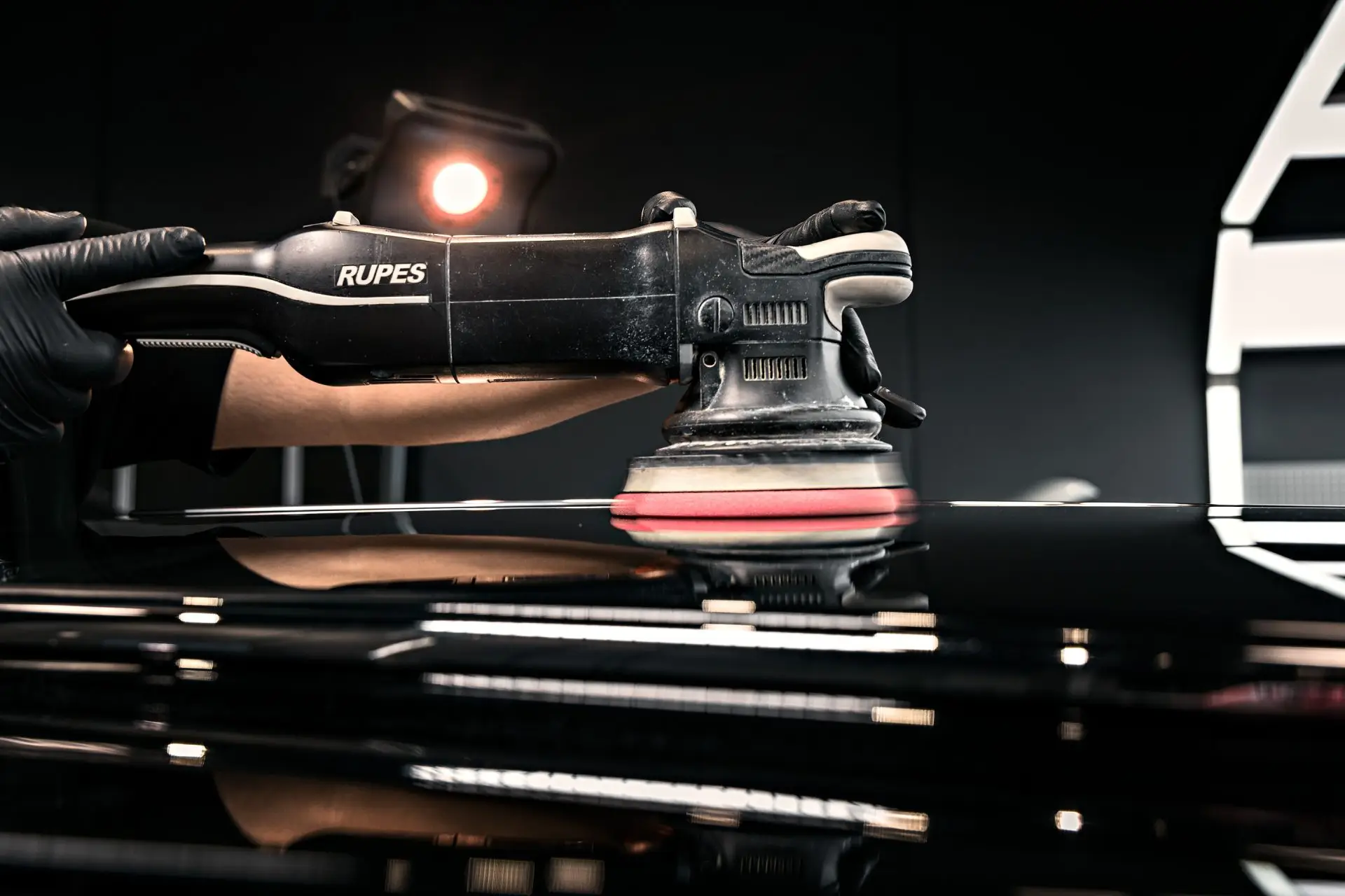 Gloved hands guiding a RUPES dual-action polisher across black paint