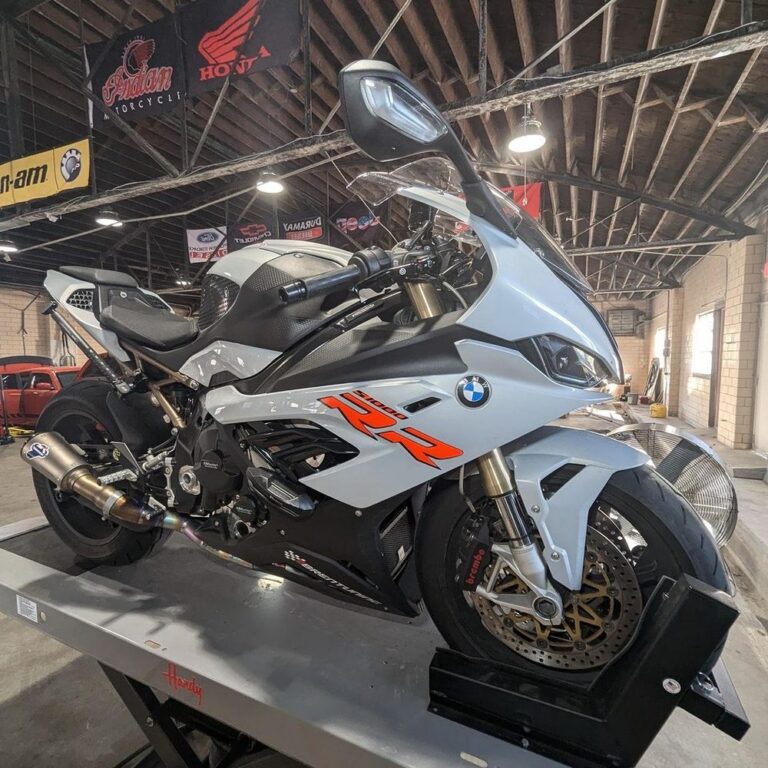 White BMW S 1000 RR superbike on a motorcycle lift in the shop, brand banners on the wall behind it