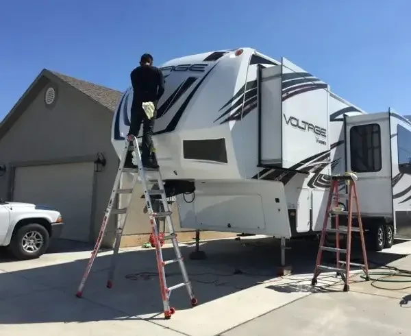 Technician on a stepladder cleaning the front cap of a Voltage toy-hauler fifth-wheel, a second ladder beside it