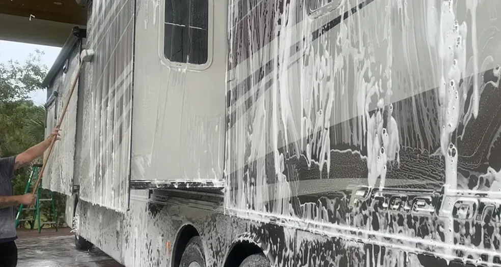 White RV covered in wash foam while a technician works it with a brush on a long pole