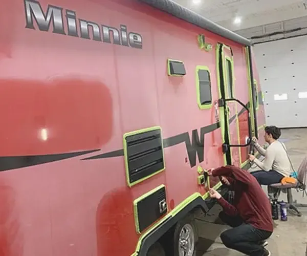 Two technicians polishing the lower side of a red Winnebago Minnie travel trailer inside a shop, decals taped off