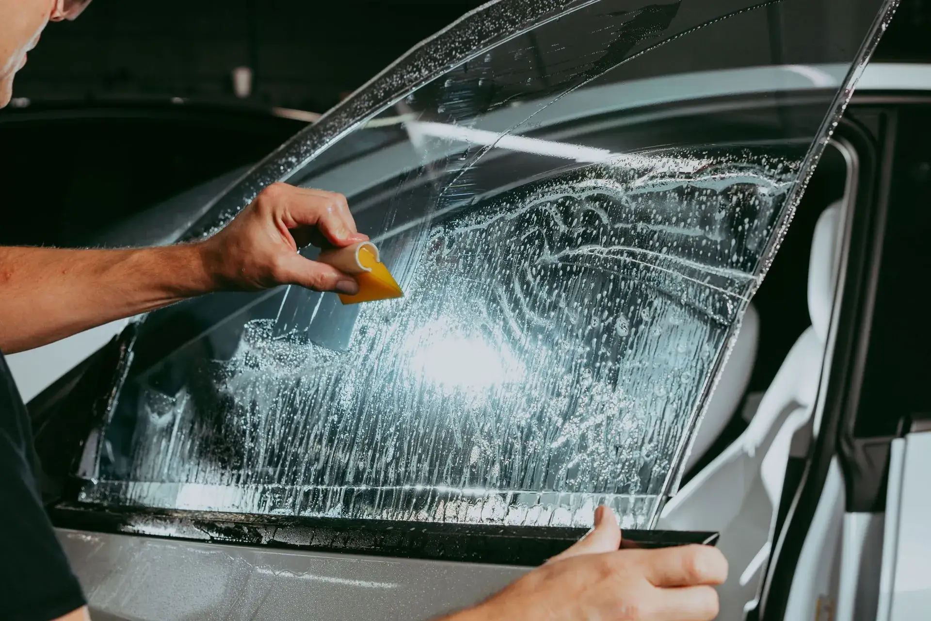 Gloved hand drawing a yellow squeegee across wet window film on a car's glass, slip solution beaded over the surface